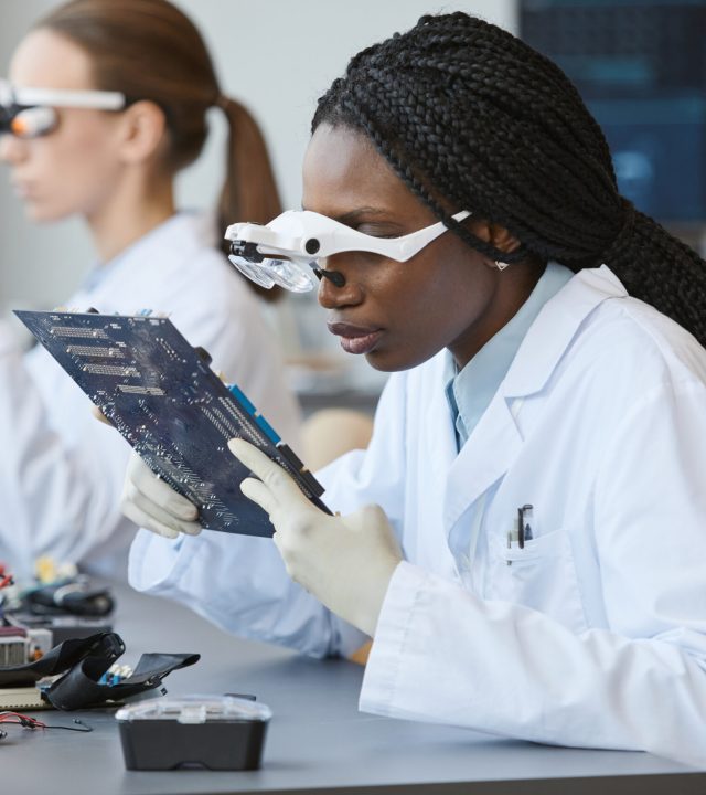 Portrait of young black woman wearing magnifying glasses and inspecting electronic parts in quality control lab